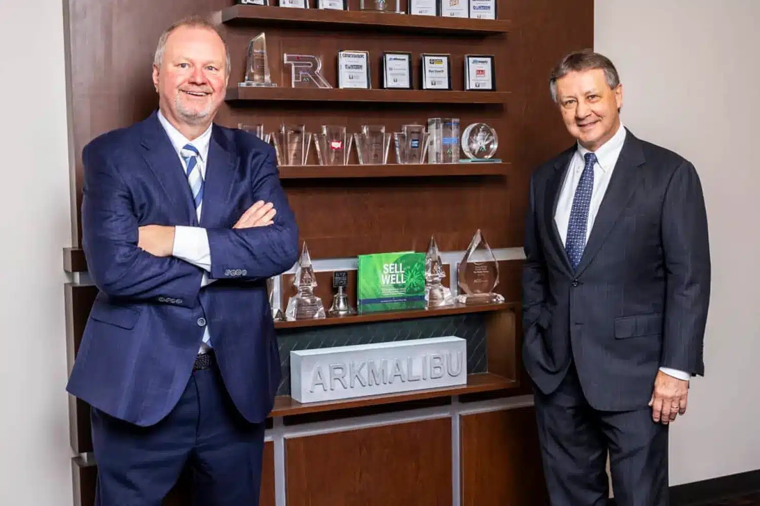 Two men in suits stand smiling in front of a wooden display shelf with trophies, awards, and a sign reading ARK MALIBU. The man on the left has arms crossed; both appear professional and confident.